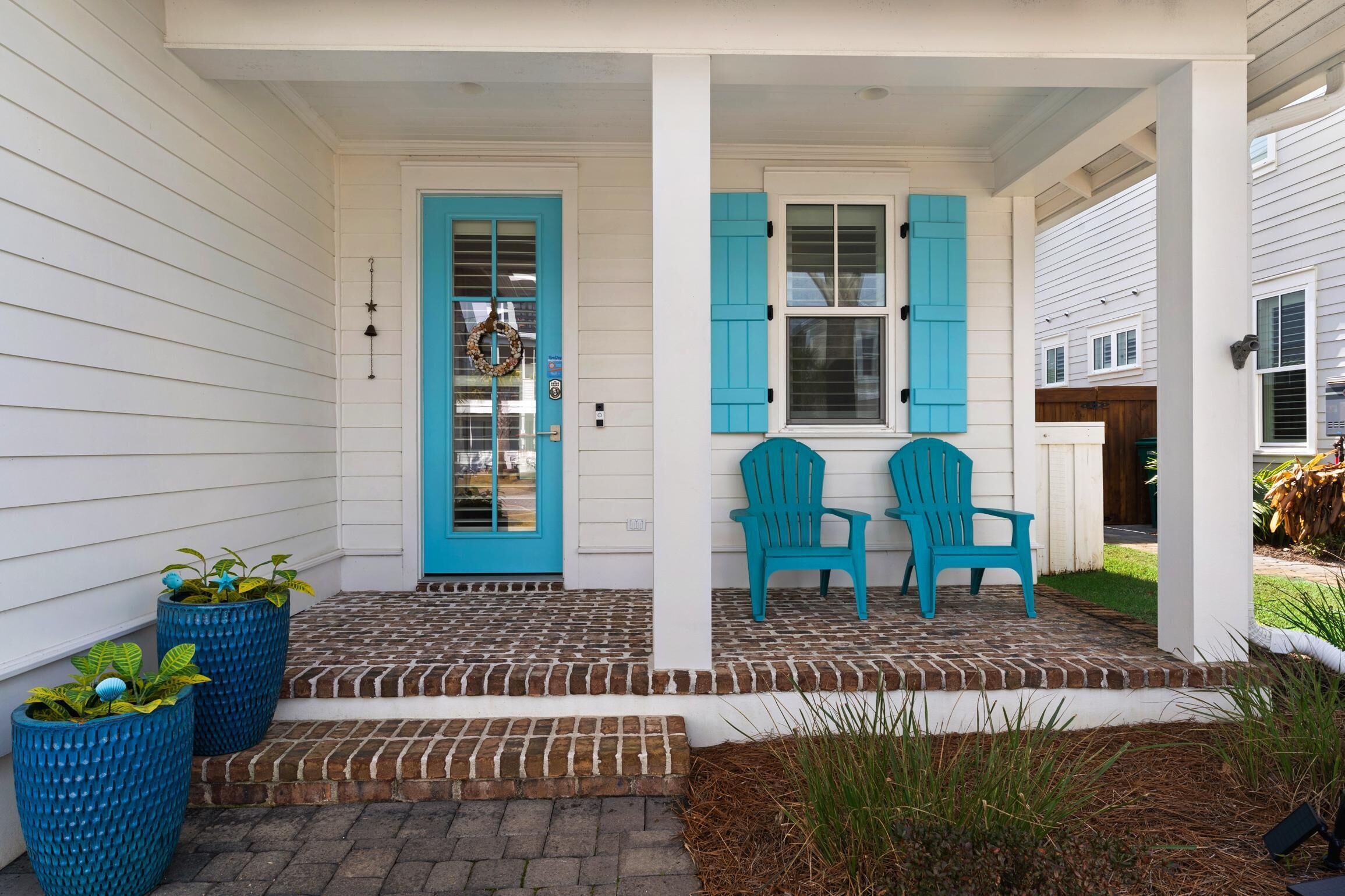 A porch with two blue Adirondack chairs, a white door with a wreath, and a brick pathway leading to the door.
