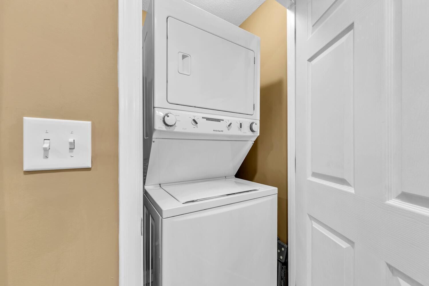 A compact washer and dryer unit installed in a corner of a room with beige walls and a white door.