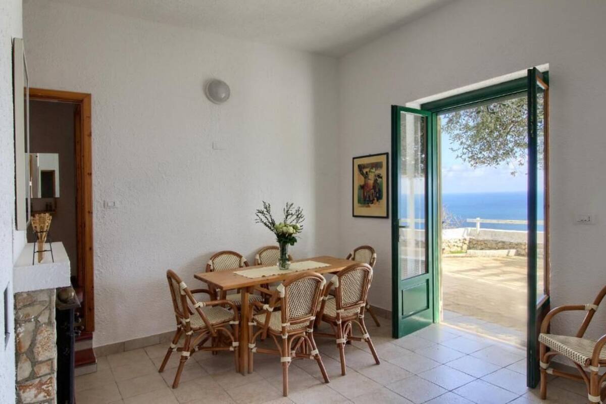 Ein Essbereich mit Holztisch und Korbstühlen, einer Blumenvase und Blick auf das Meer durch eine Glastür.