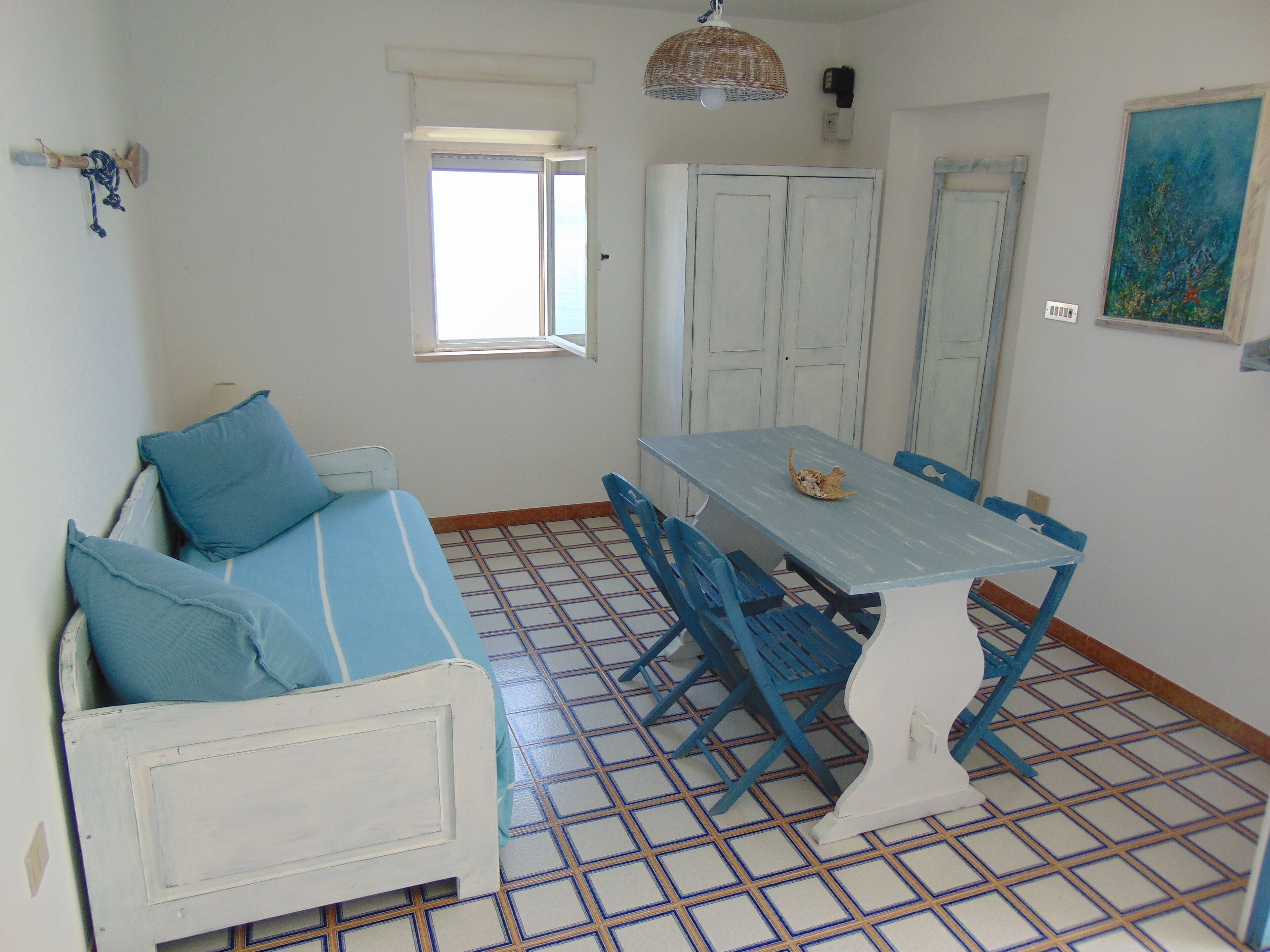 Una habitación con un sofá blanco y cojines azules, una mesa azul con sillas, una ventana, una puerta y un cuadro en la pared.