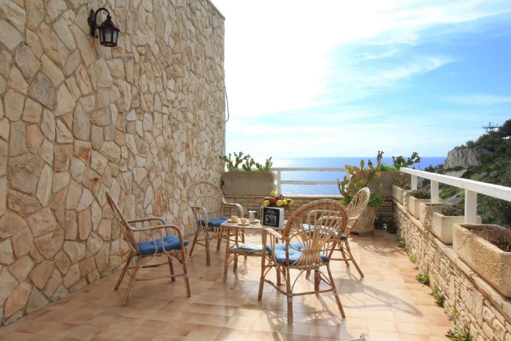 A stone-walled outdoor seating area with wicker chairs and a table, overlooking a coastal view.