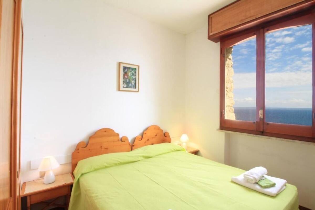 Ein Schlafzimmer mit Bett, Nachttisch, Lampe, Fenster mit Meerblick und einem gerahmten Bild an der Wand.