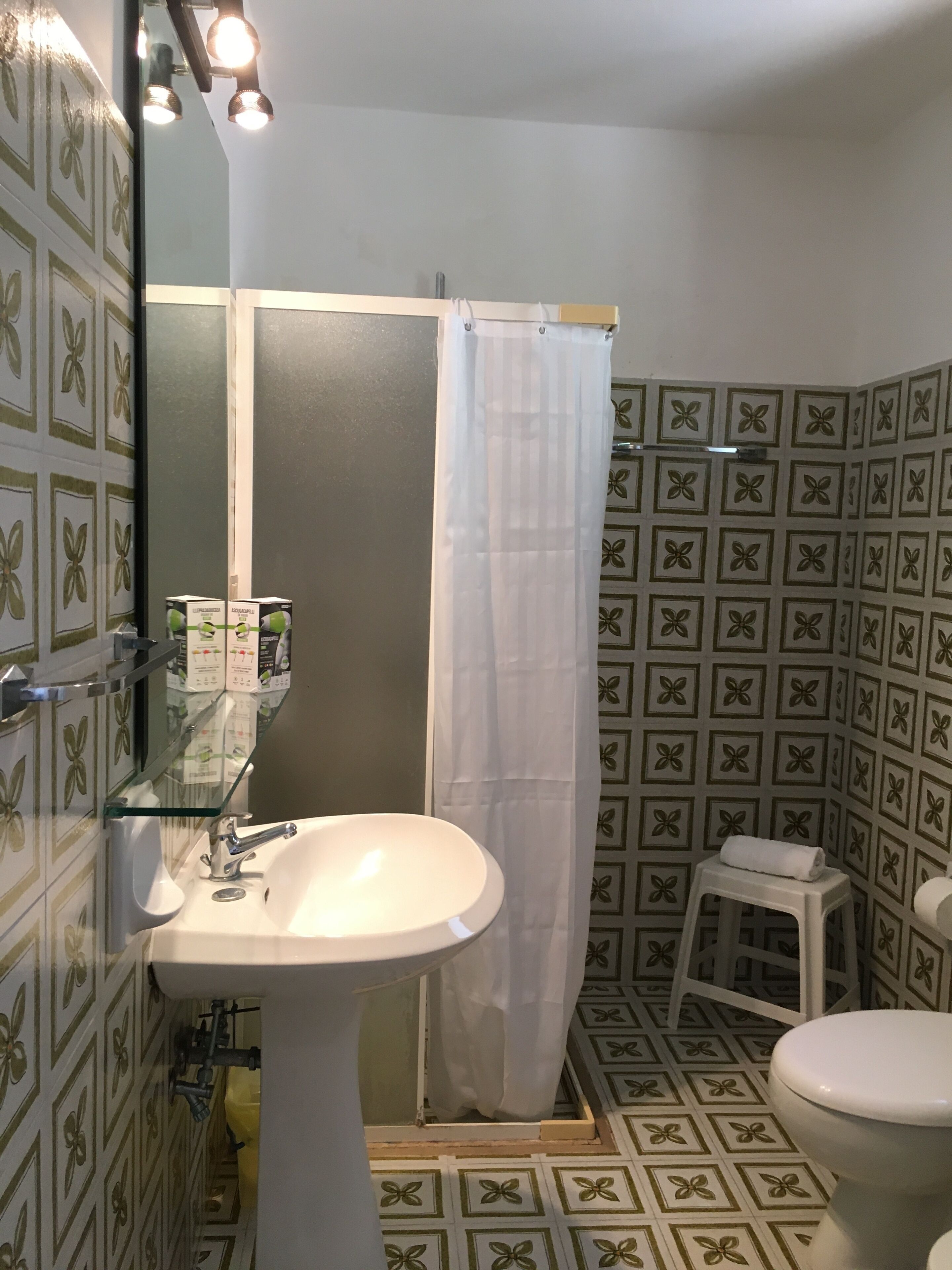 Un baño con lavamanos blanco, repisa de vidrio, cortina de ducha, inodoro y paredes con diseño.