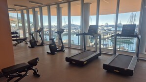 Un gimnasio con cintas de correr, elípticas y un banco, con vistas a una marina con barcos y yates.