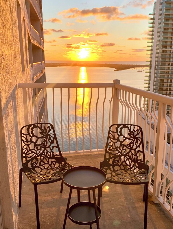 Un balcone con due sedie e un tavolino, affacciato su un tramonto su un corso d'acqua con un ponte all'orizzonte.