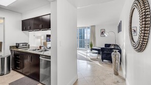 A modern kitchen with dark cabinets and a built-in oven, a living area with a dark sofa and a decorative vase, and a large window offering a city view.