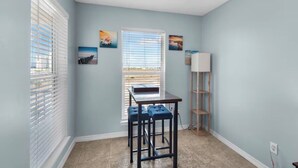 A room with light blue walls, a window with blinds, a desk with a chair, and a shelf unit. - Sunset Suite (Navarre)