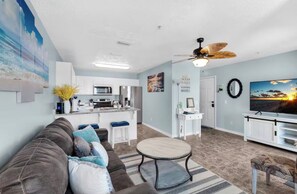 A living room with a sofa, a coffee table, and a TV. There is a kitchen area with cabinets and appliances.