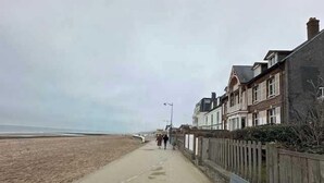 Een kustpad met een bankje, naast een zandstrand en omzoomd door woongebouwen.