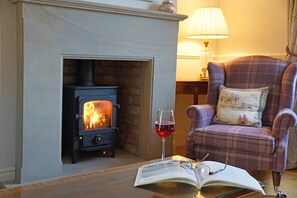 A room with a fireplace, a plaid armchair, a glass of red wine, and an open book on a table.