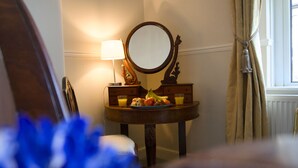 Ein Zimmer mit einem runden Spiegel, einem kleinen Tisch mit Obst und Saft, einer Lampe und einem Fenster mit Vorhängen.