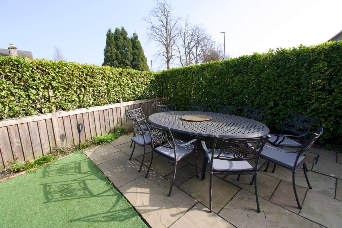 A round metal table with six chairs, surrounded by a wooden fence and a hedge.