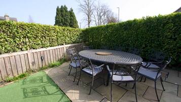 A round metal table with six chairs, surrounded by a wooden fence and a hedge.