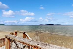 Una terraza de madera con vista a una playa y un mar tranquilo, con tierra a lo lejos.