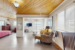 Un salón espacioso con suelo de madera, un sofá, un sillón, un televisor y un ventanal grande con persianas.