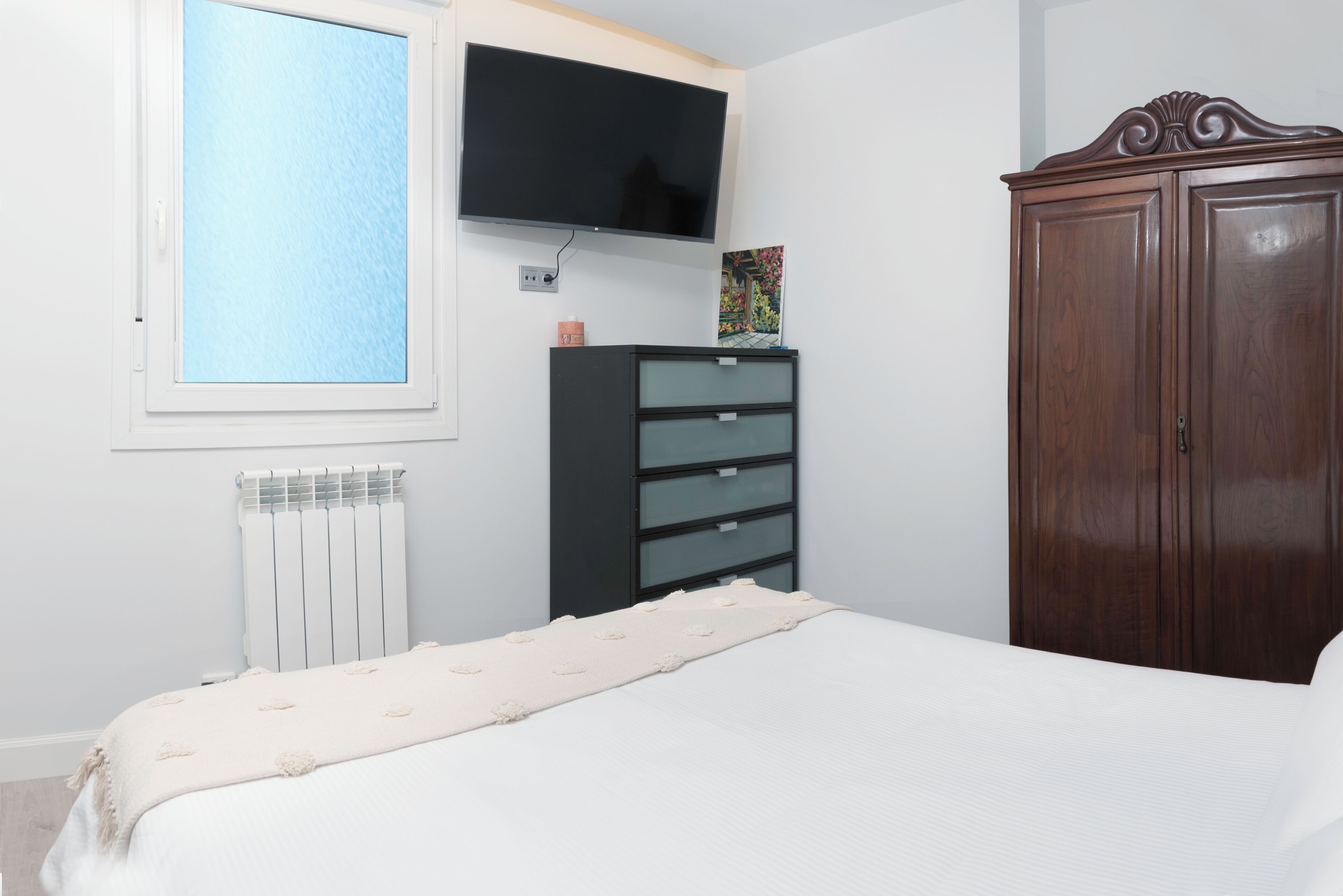 Un dormitorio con una cama, un aparador, un televisor montado en la pared y un radiador.