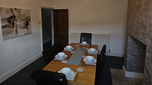 Une salle à manger avec une longue table en bois dressée pour quatre personnes, des chaises, un lustre moderne, un tableau au mur et une cheminée.