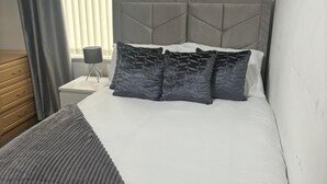 Un lit bien fait, avec une tête de lit grise, une table de chevet avec une lampe et une fenêtre avec des rideaux.