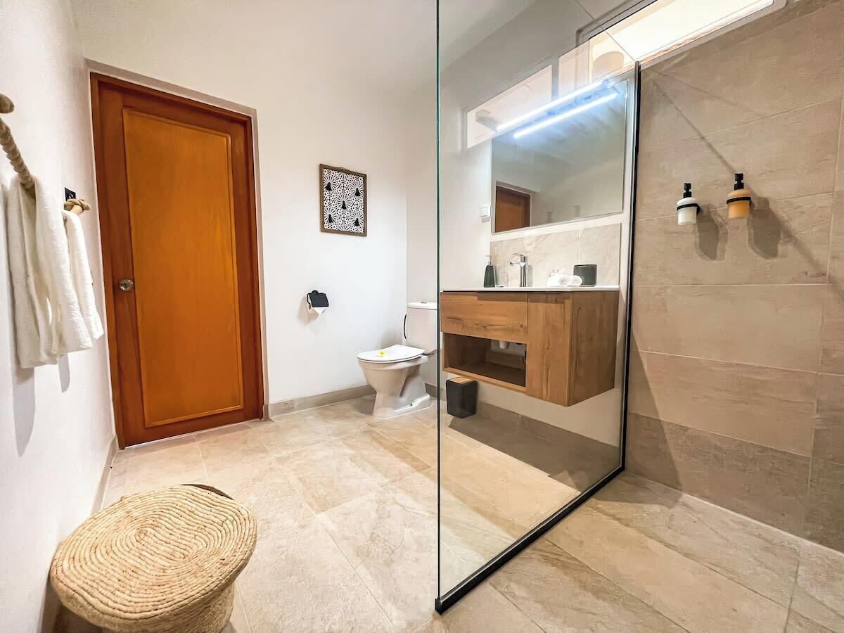 Ein Badezimmer mit Holztür, Toilette, Waschbecken mit Spiegel und einem gewebten Hocker.