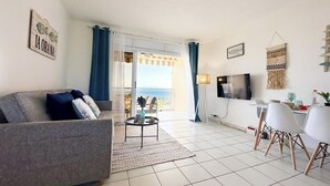 Ein modernes Wohnzimmer mit grauem Sofa, einem kleinen Couchtisch, einem Essbereich mit weißen Stühlen, einem an der Wand befestigten Fernseher und einem Balkon mit blauen Vorhängen.