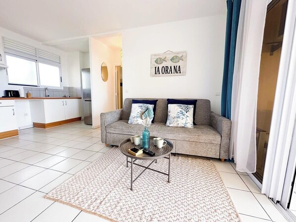 A living room with a sofa, coffee table, and a wall sign reading "IA ORANA".
