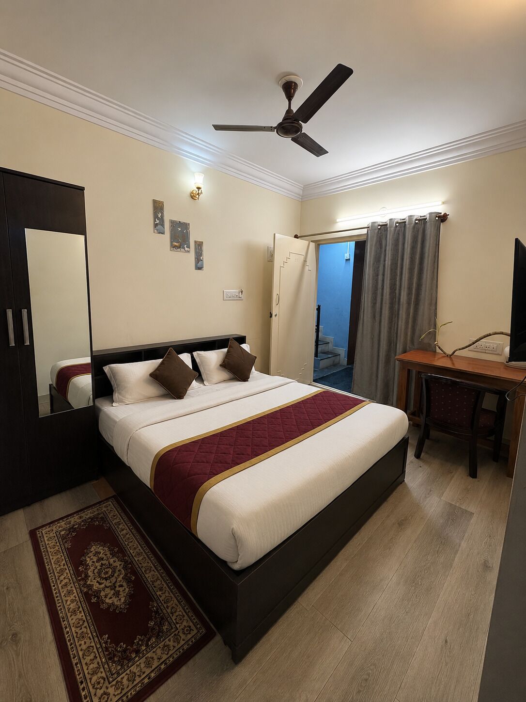Habitación de hotel con cama, escritorio, ventilador de techo, espejo y puerta que da a un balcón.