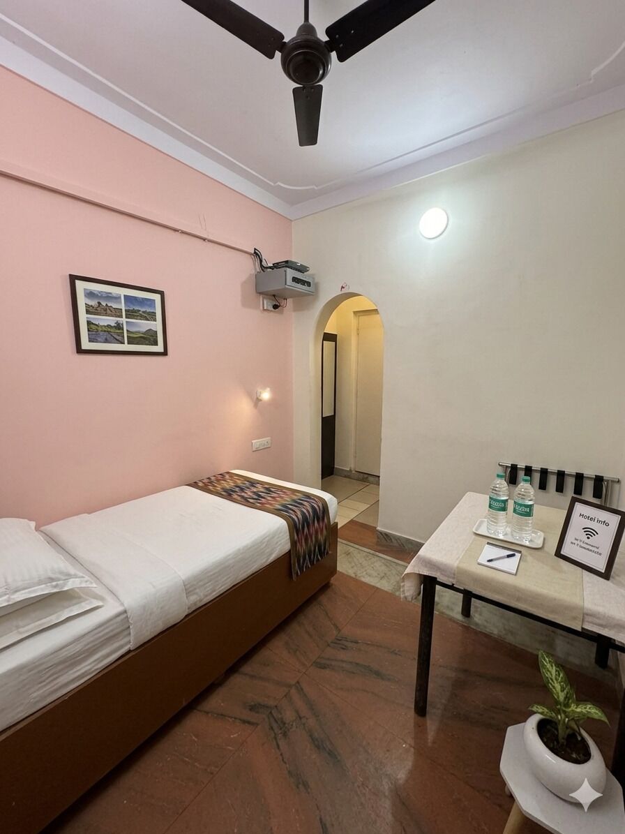 A single bed with a headboard, a small table with a plant, a ceiling fan, and a framed picture on the wall.