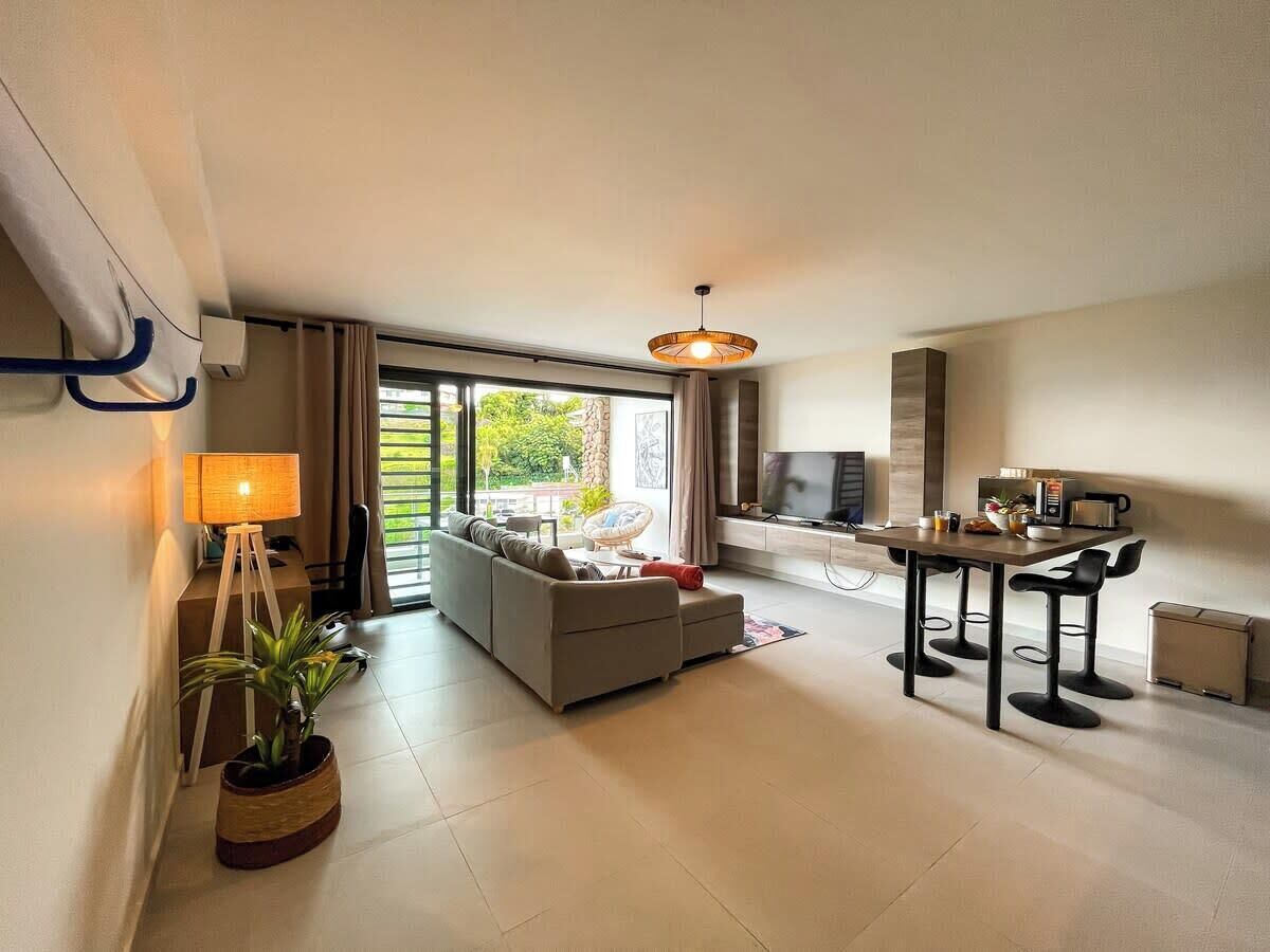 A modern living room with a sofa, a dining table with bar stools, a TV, and a large window with curtains.