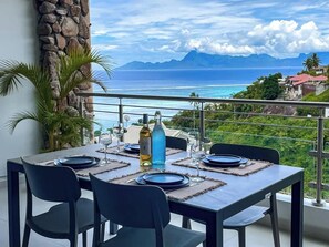 Mesa de jantar posta com pratos, talheres e copos de vinho, com vista para a costa, montanhas e céu claro.