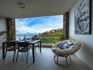 Uma área de refeições com vista para o mar e montanhas, uma cadeira de vime e uma mesa posta para dois.