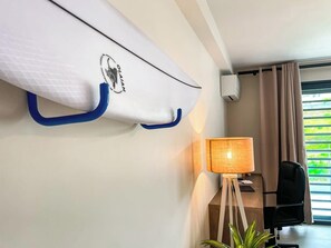 Quarto de hotel com ar condicionado fixado na parede, luminária sobre pedestal de madeira, cadeira e vista para o verde através de uma janela com cortinas.