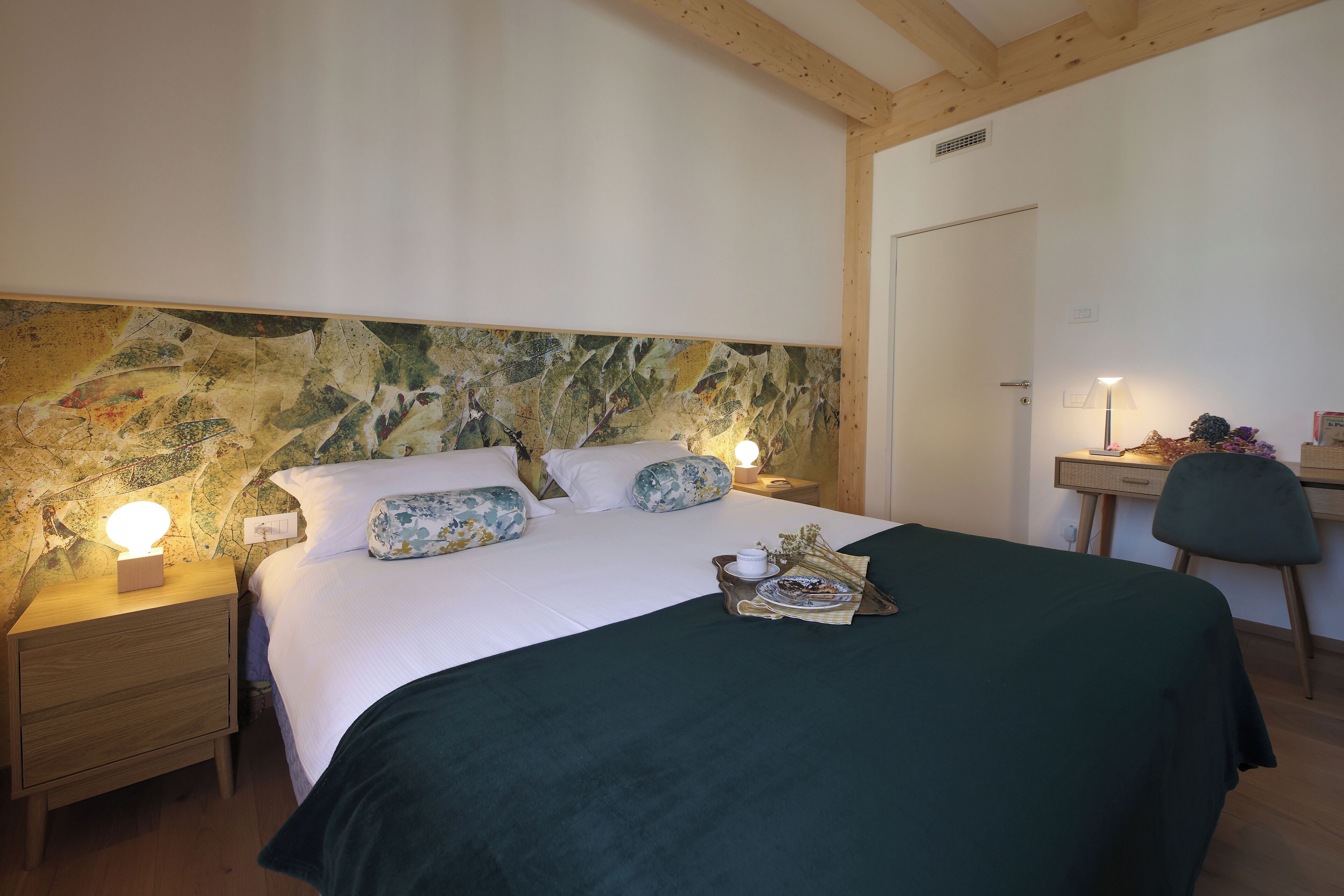 Un lit bien fait, avec une couvre-lit verte et des coussins à motifs floraux, une table de chevet avec une lampe, et un bureau avec une chaise, dans une pièce aux poutres apparentes et dont la tête de lit est décorée d’un motif en pierre.