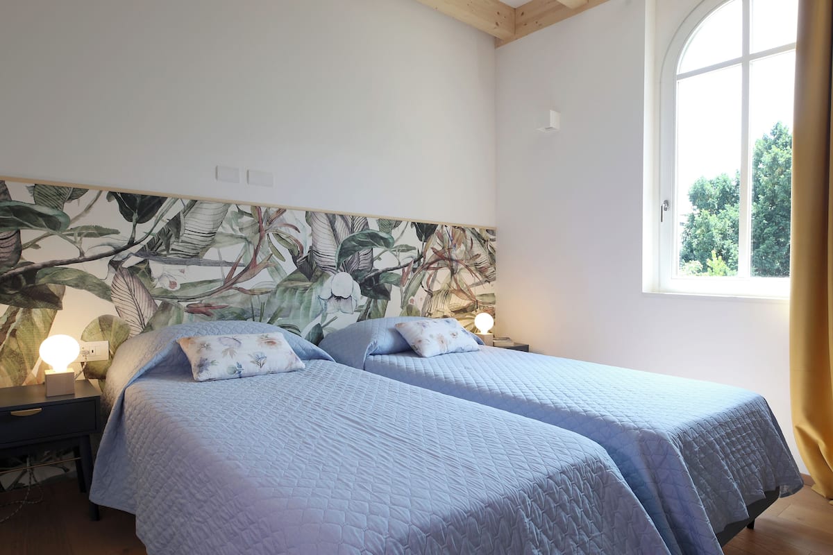 A bedroom with two beds, a wooden ceiling, a large window, and a wall with a botanical mural.