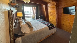 Une chambre confortable, aménagée dans une cabane en bois, avec un lit, un luminaire suspendu, un téléviseur fixé au mur et une vue sur l’extérieur par une fenêtre.