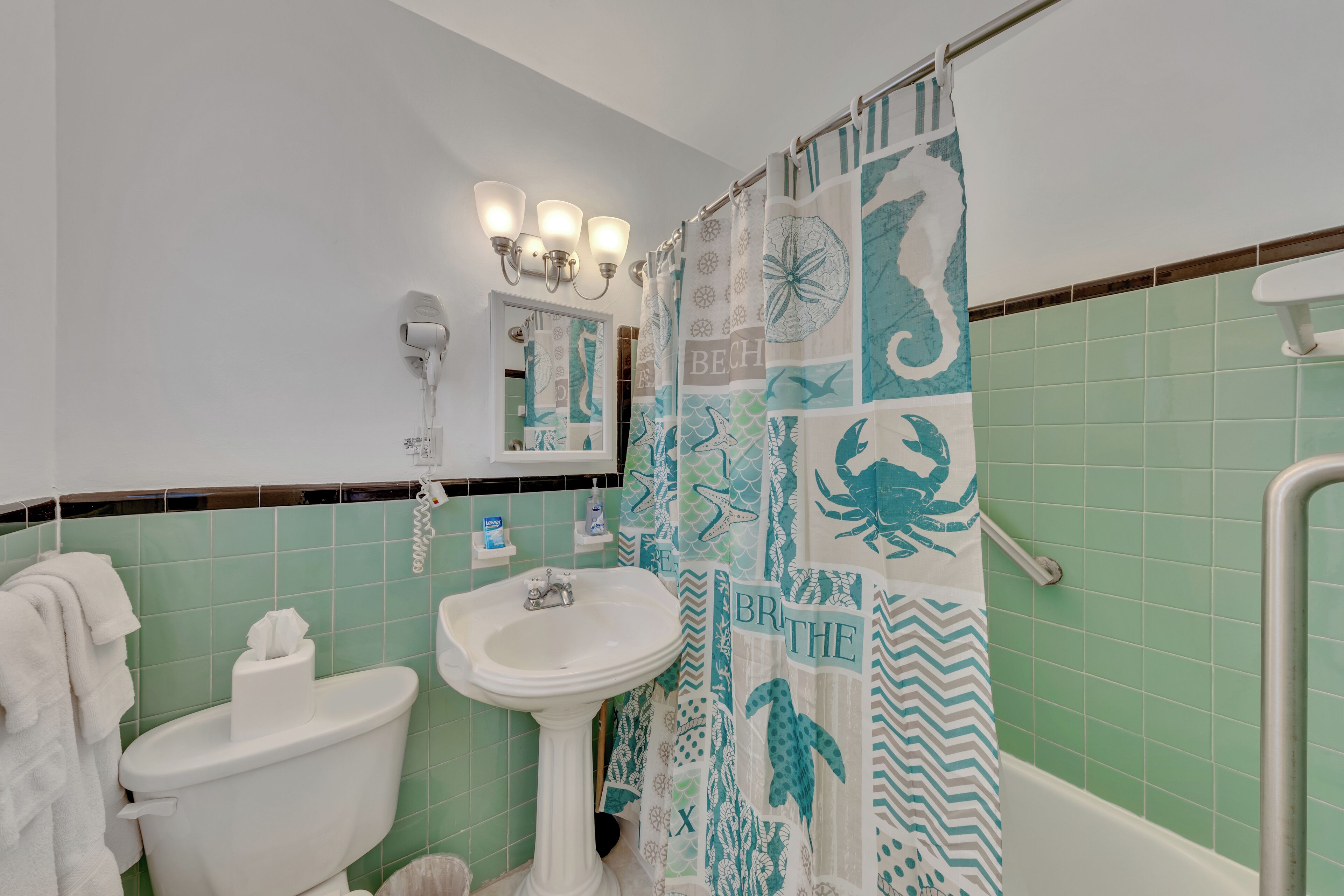 Un baño con inodoro blanco, lavamanos pedestal y cortina de ducha con diseño de playa.