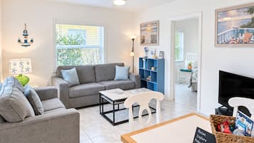 A modern living room with a grey sofa, a coffee table, and a TV. There is a bookshelf, a wicker basket, and a sign that reads 'Relax Explore Make Memories Welcome!'