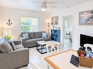 A modern living room with a grey sofa, a coffee table, and a TV. There is a bookshelf, a wicker basket, and a sign that reads 'Relax Explore Make Memories Welcome!' - Bahama Beach Club Pompano (Pompano Beach)