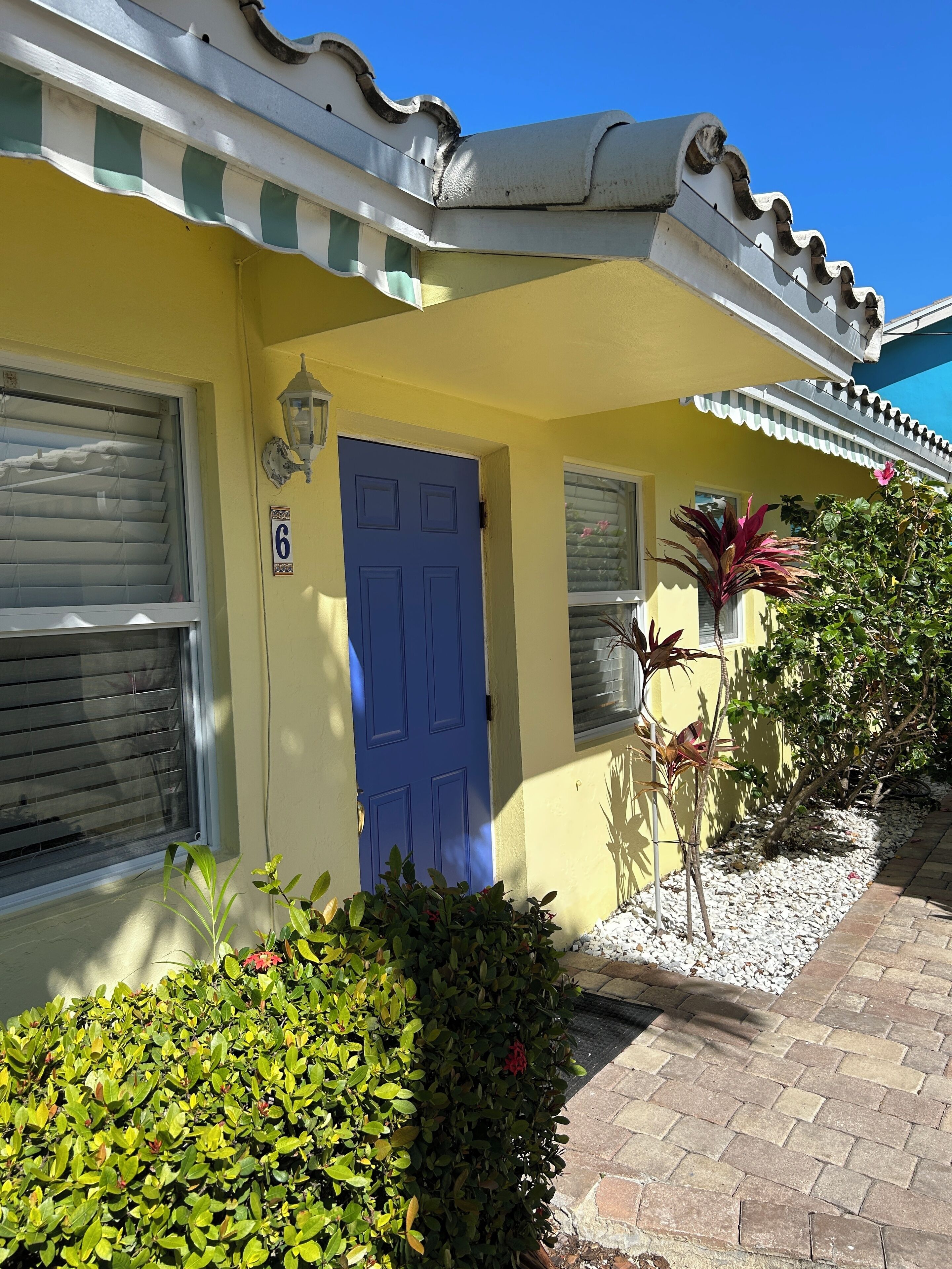 A yellow building with a blue door, a window with shutters, and a small garden with plants and a pathway.