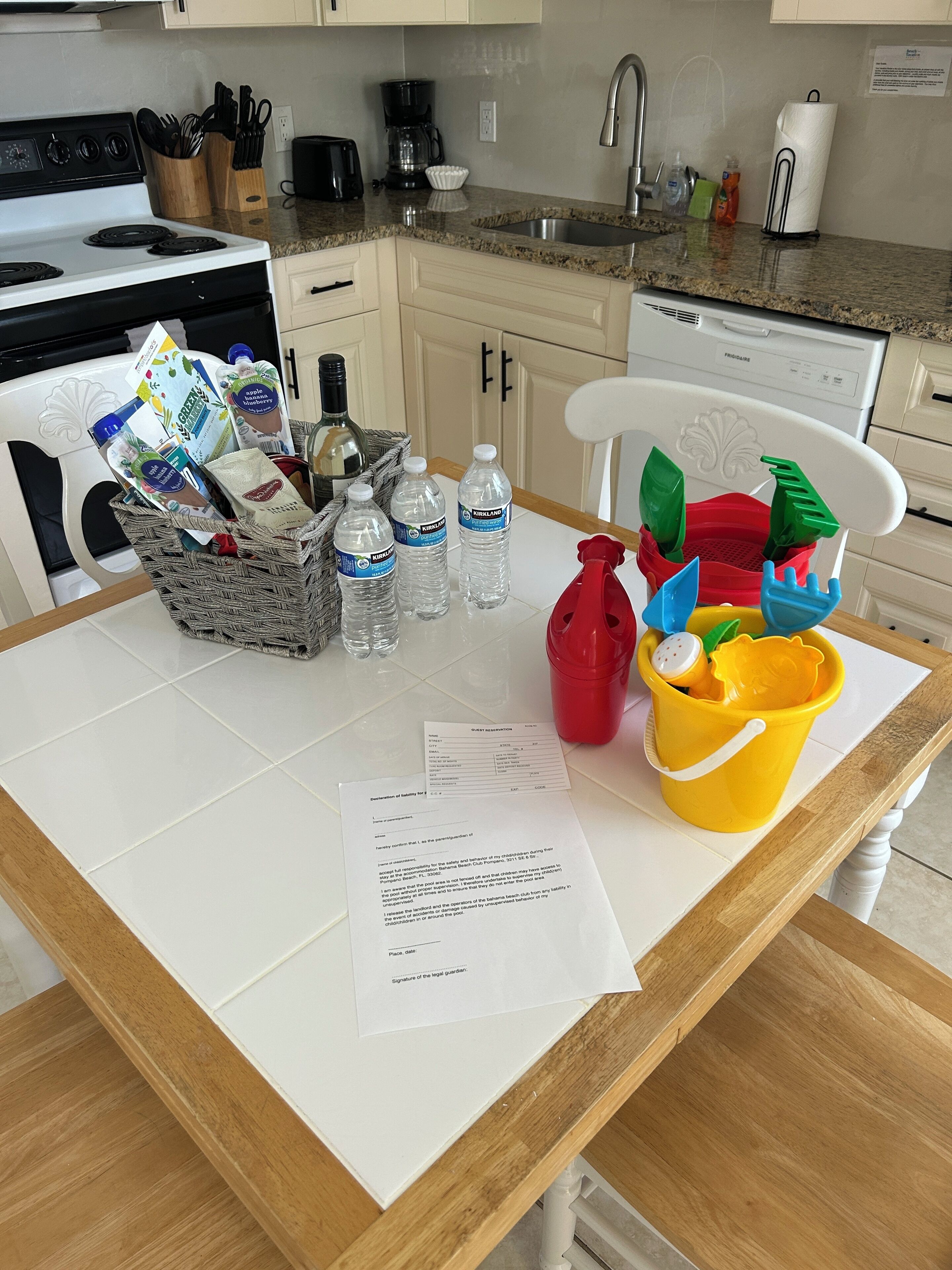 Une cuisine dotée d’un plan de travail en granit, de meubles blancs, d’une cuisinière et d’un lave-vaisselle. On trouve divers objets sur le plan de travail, notamment un panier, des bouteilles d’eau, un vaporisateur rouge et un seau jaune rempli de jouets.
