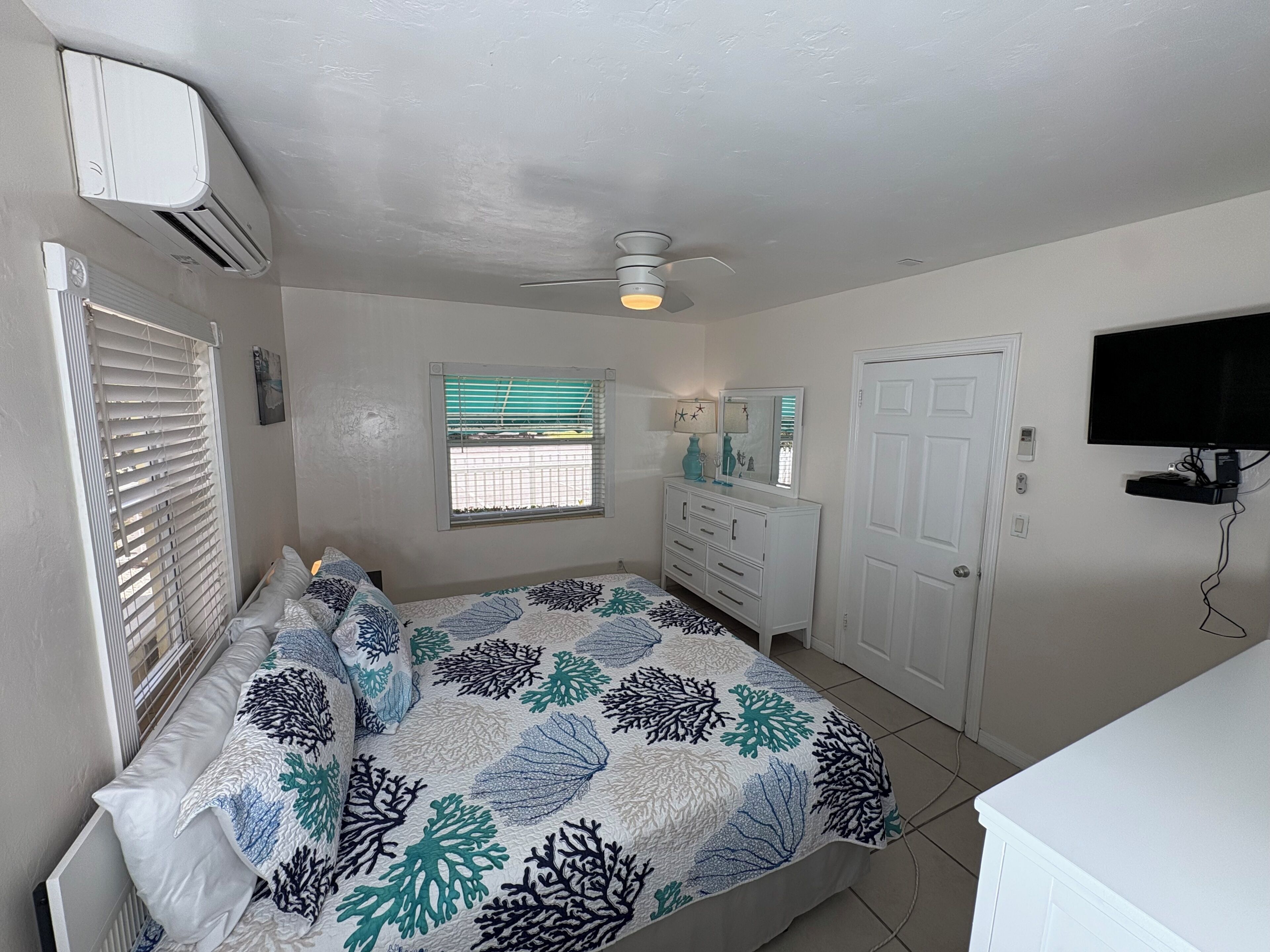 Quarto com cama, penteadeira, ventoinha no teto, janela com persianas, porta e televisão fixada na parede.