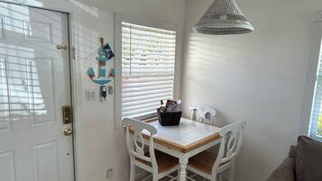 Un coin repas avec une table et des chaises blanches, un luminaire suspendu et une fenêtre avec des stores.