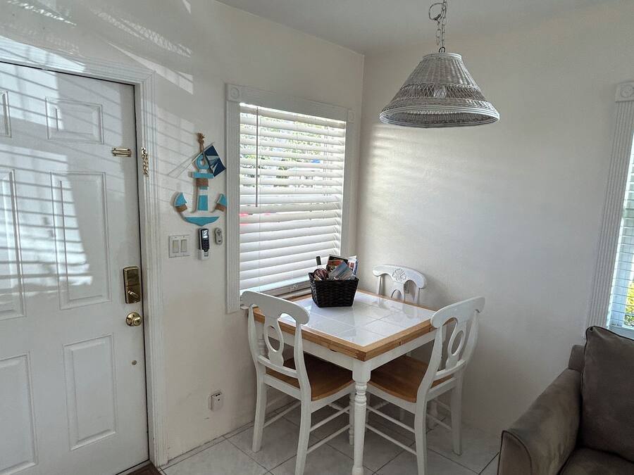 Un coin repas avec une table et des chaises blanches, un luminaire suspendu et une fenêtre avec des stores.
