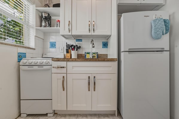 A kitchen with white cabinets, a stove, a refrigerator, and a window with blinds. - Bahama Beach Club Pompano (Pompano Beach)