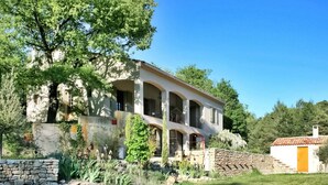 A large, multi-story building with arched windows and a stone facade, surrounded by lush greenery and a stone wall. - La Caracole :Provencal villa with heated pool, tennis court, nature at your doorstep (Caseneuve)