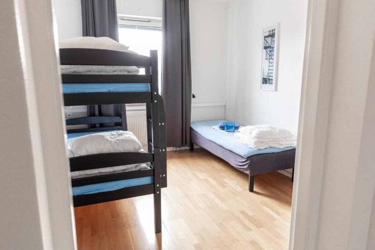Une chambre de dortoir avec des lits superposés et un lit simple, un plancher en bois, une fenêtre avec des rideaux et un tableau encadré au mur.