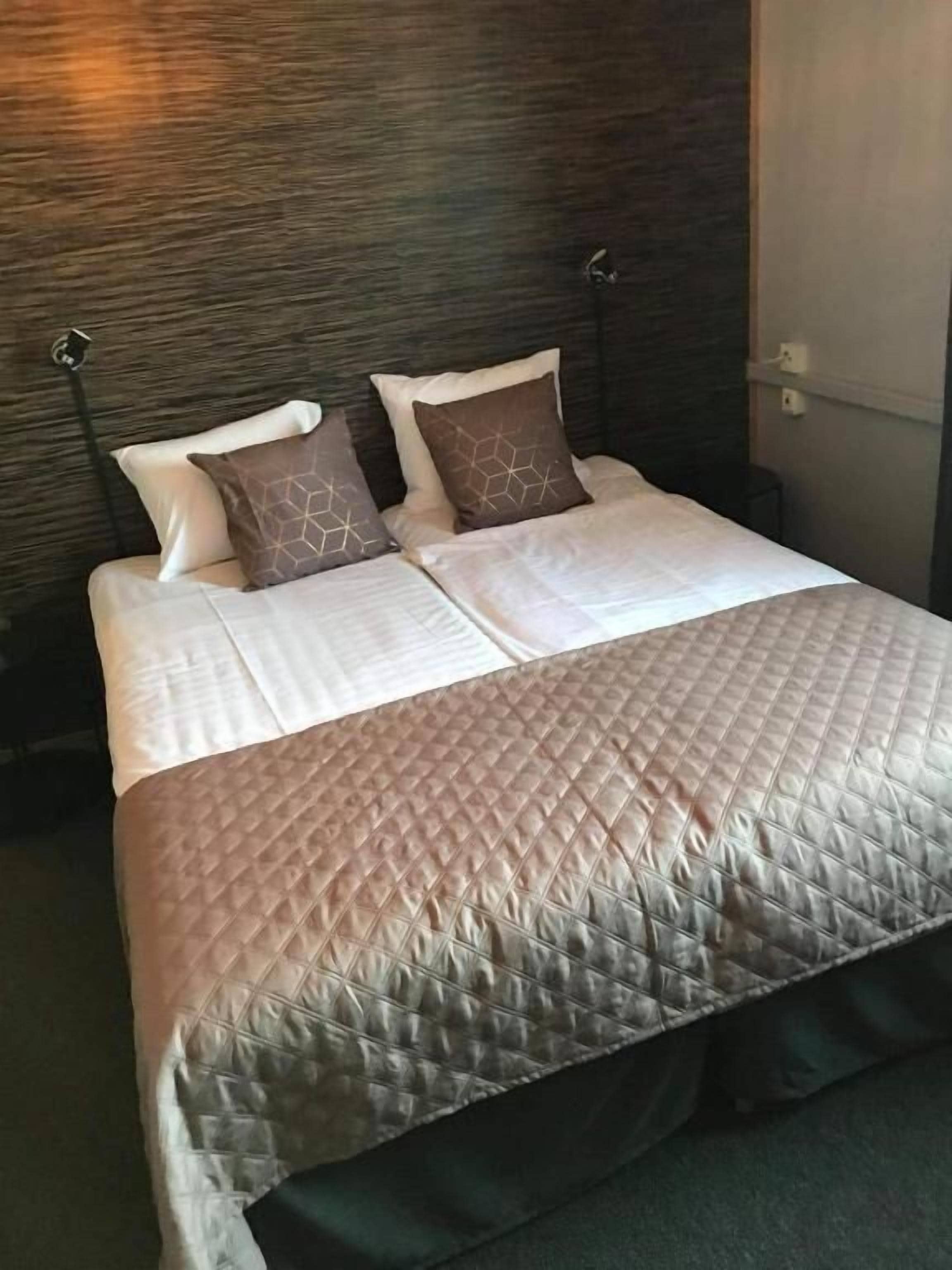 Una cama bien hecha con cubrecolcha acolchado, dos almohadas y cabecera frente a una pared con textura.