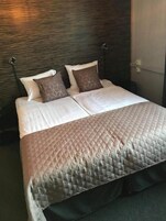 Una cama bien hecha con cubrecolcha acolchado, dos almohadas y cabecera frente a una pared con textura.