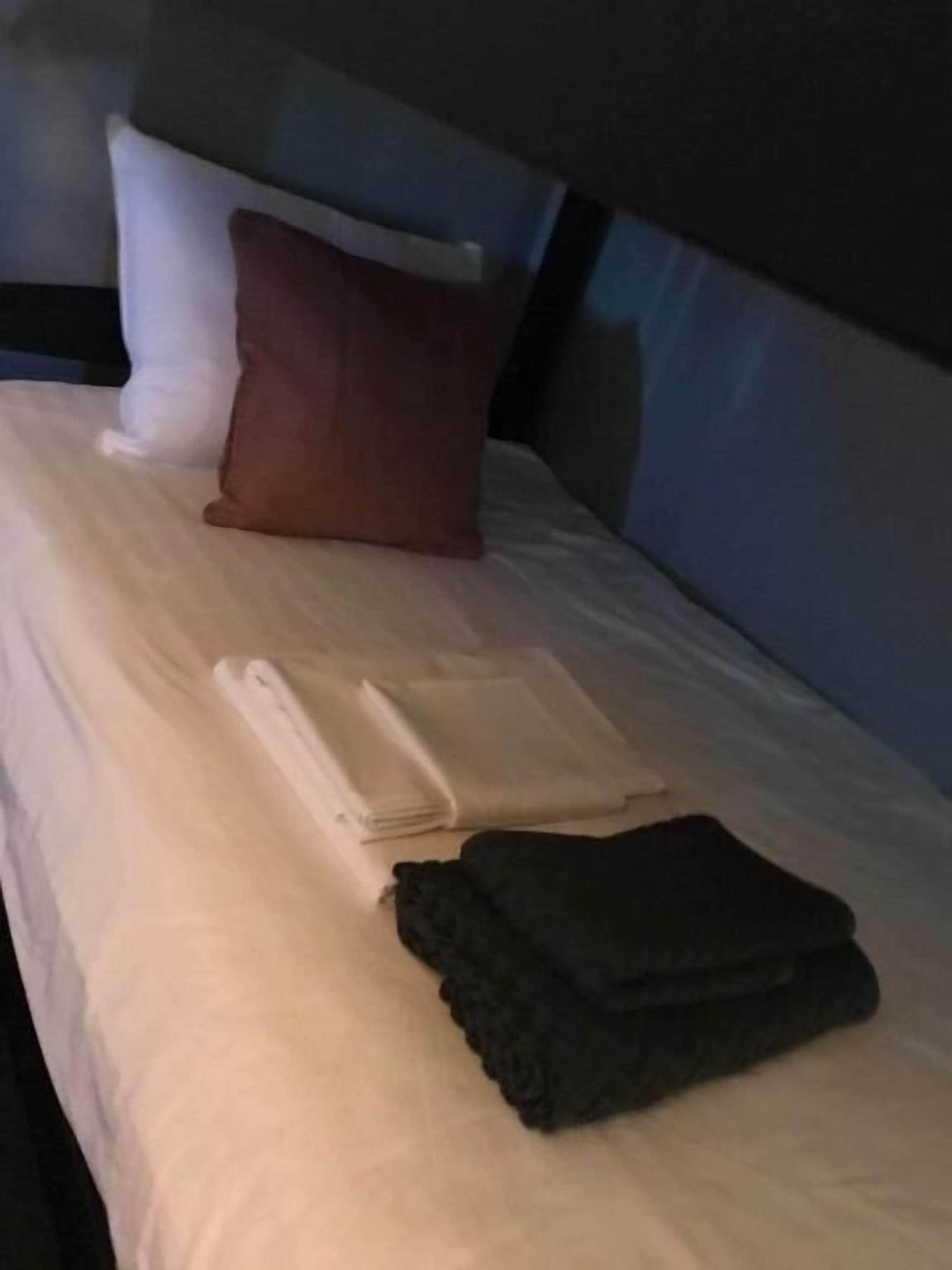 Una cama bien hecha con una almohada blanca, una almohada marrón, una toalla blanca doblada y una manta oscura doblada.