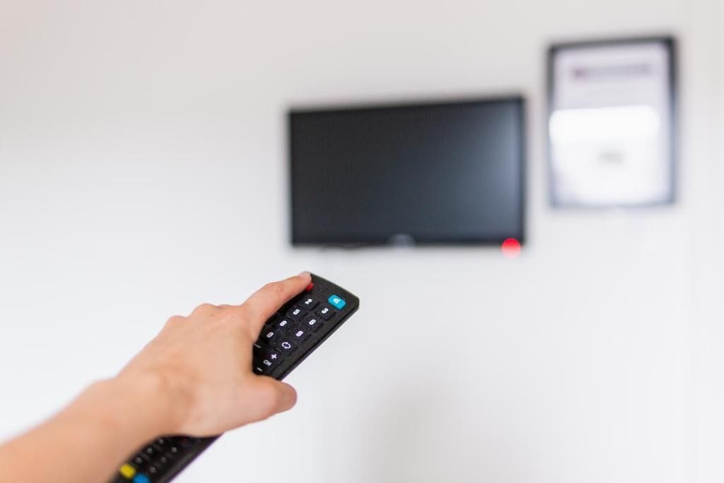A hand holding a remote control in front of a television and a wall-mounted device.
