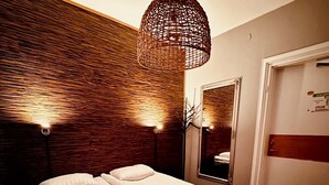 Cama bem arrumada com lençóis brancos e uma toalha dobrada, luminária de tecido acima e um espelho na parede.